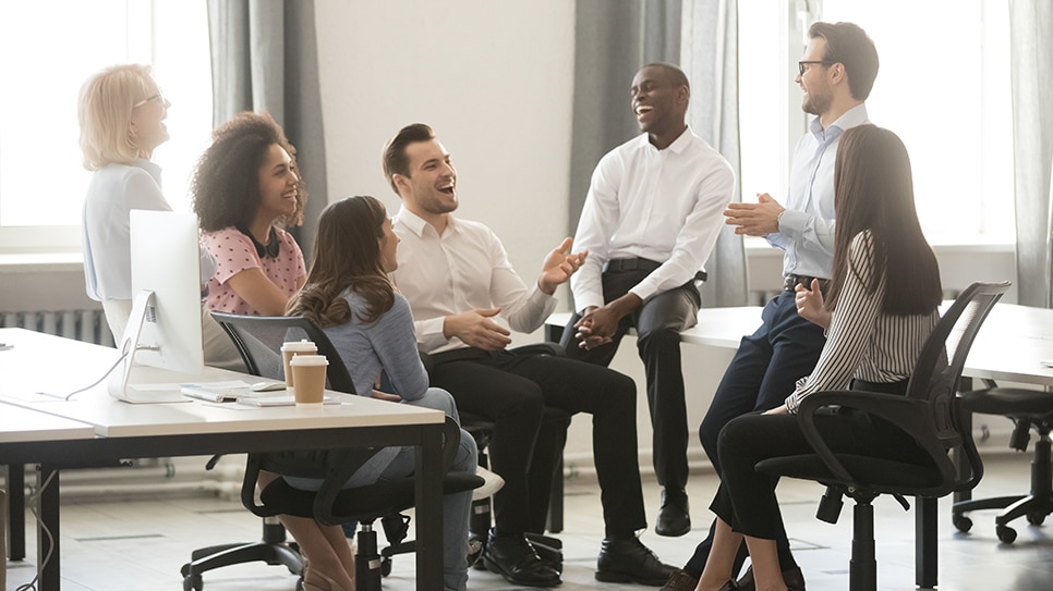  A group of diverse employees are sitting and standing in a circle in the office, talking and laughing, with the intent to evaluate and improve their work environment.